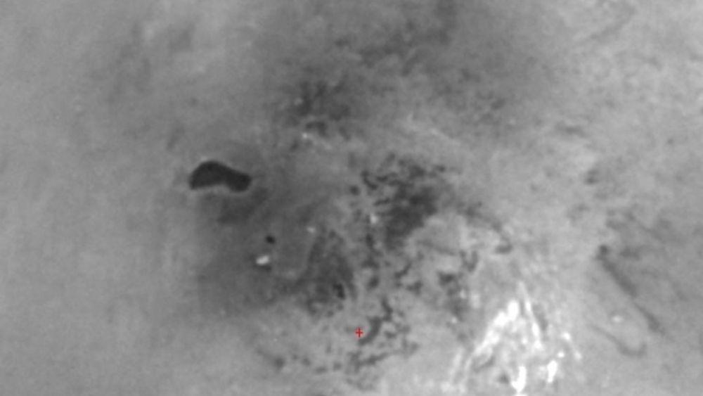 SYDPOL: Bildet viser Titans sydpol (rødt kryss -  se stor versjon av bildet). Det skarpt avgrensede, mørke området kan tenkes å være en metansjø. Titan er isende kald og metan forekommer bare flytende. Eventuelt vann forekommer bare som is.